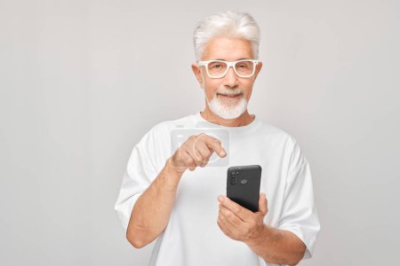 Téléchargez les photos : Portrait d'homme aux cheveux gris en T-shirt blanc montrant écran de téléphone portable vierge avec visage excité. Personne avec smartphone isolé sur fond blanc - en image libre de droit