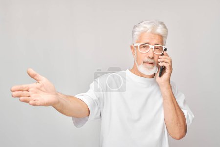 Foto de Retrato del hombre de pelo gris en camiseta blanca hablando por teléfono móvil. Persona con teléfono inteligente aislado en el fondo blanco - Imagen libre de derechos