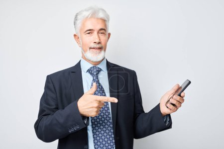 Téléchargez les photos : Portrait d'un homme d'affaires âgé en costume montrant un écran vierge de téléphone portable avec un visage excité. Personne avec smartphone isolé sur fond blanc - en image libre de droit