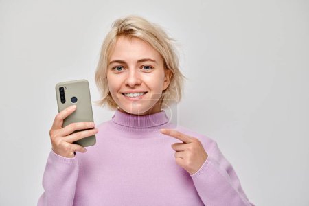Foto de Retrato de una joven rubia sosteniendo el teléfono móvil en la mano con la cara sonriente feliz. Persona con teléfono inteligente aislado en el fondo blanco - Imagen libre de derechos