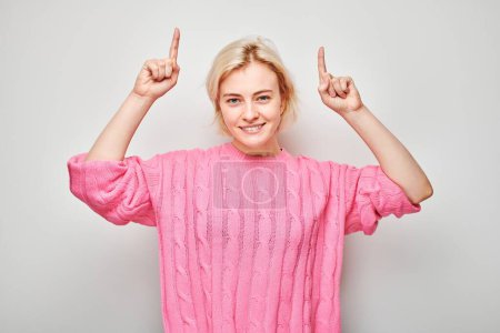 Téléchargez les photos : Jeune femme blonde en pull rose pointant le doigt vers le haut, démontrant un espace vide pour le produit ou le texte isolé sur fond de studio blanc - en image libre de droit