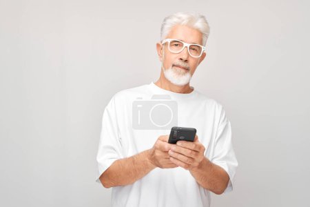 Téléchargez les photos : Portrait d'homme aux cheveux gris en T-shirt blanc tenant le téléphone portable à la main avec un visage souriant heureux. Personne avec smartphone isolé sur fond blanc - en image libre de droit