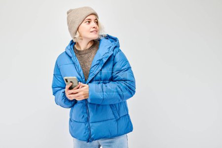 Téléchargez les photos : Portrait de jeune femme blonde vêtue d'une veste bleue tenant un téléphone portable à la main avec un visage souriant heureux. Personne avec smartphone isolé sur fond blanc - en image libre de droit
