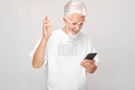 Foto de Retrato de hombre de pelo gris en camiseta blanca sosteniendo el teléfono móvil en la mano con la cara sonriente feliz. Persona con teléfono inteligente aislado en el fondo blanco - Imagen libre de derechos