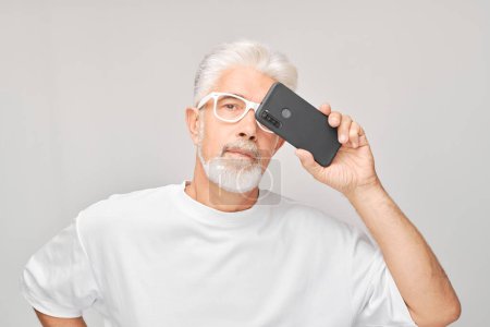 Foto de Retrato de hombre de pelo gris en camiseta blanca toma foto selfie en el teléfono móvil. Blogger con smartphone aislado sobre fondo blanco - Imagen libre de derechos