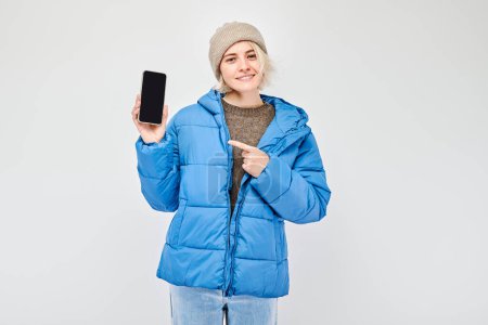Foto de Retrato de una joven rubia con chaqueta azul que muestra la pantalla del teléfono móvil en blanco con la cara excitada. Persona con teléfono inteligente aislado en el fondo blanco - Imagen libre de derechos