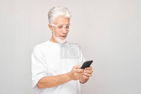 Photo for Portrait of gray-haired man in white T-shirt looks on mobile phone and thinks. Person with smartphone isolated on white backgroun - Royalty Free Image