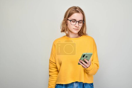 Téléchargez les photos : Portrait de jeune femme blonde regarde sur le téléphone portable et pense. Personne avec smartphone isolé sur fond blanc - en image libre de droit
