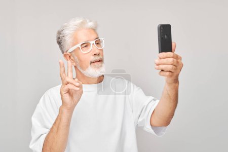 Foto de Retrato de hombre de pelo gris en camiseta blanca toma foto selfie en el teléfono móvil. Blogger con smartphone aislado sobre fondo blanco - Imagen libre de derechos