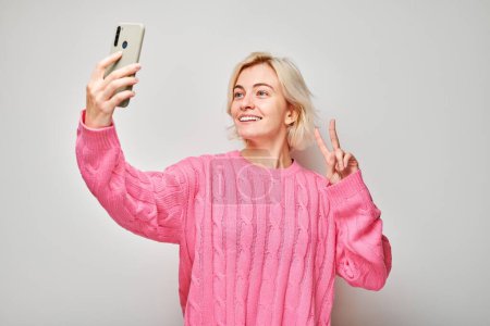 Photo for Portrait of young blond woman in pink sweater takes selfie photo on mobile phone. Blogger with smartphone isolated on white backgroun - Royalty Free Image