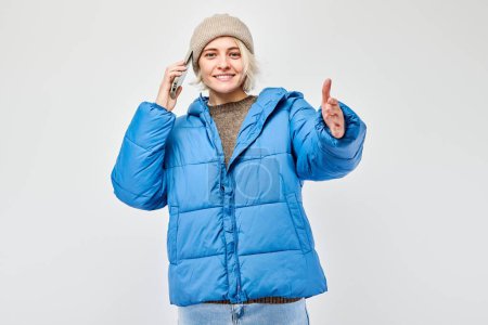 Foto de Retrato de una joven rubia con chaqueta azul hablando por teléfono móvil con cara feliz y sonriente. Persona con teléfono inteligente aislado en el fondo blanco - Imagen libre de derechos