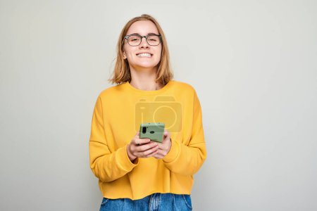 Foto de Retrato de una joven rubia sosteniendo el teléfono móvil en la mano con la cara sonriente feliz. Persona con teléfono inteligente aislado en el fondo blanco - Imagen libre de derechos