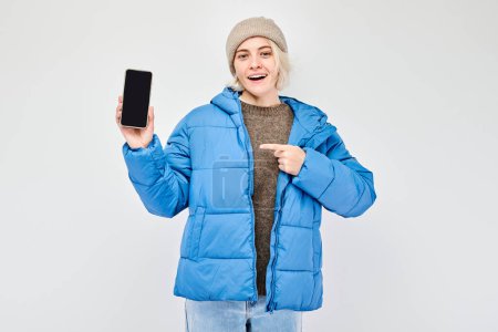 Téléchargez les photos : Portrait de jeune femme blonde veste bleue montrant écran de téléphone portable vierge avec visage excité. Personne avec smartphone isolé sur fond blanc - en image libre de droit