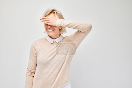 Téléchargez les photos : Portrait de jeune femme blonde couvrant les yeux à la main isolé sur fond de studio blanc - en image libre de droit