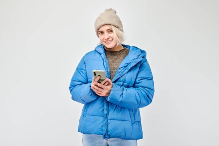 Foto de Retrato de una joven rubia con chaqueta azul sosteniendo el teléfono móvil en la mano con la cara sonriente feliz. Persona con teléfono inteligente aislado en el fondo blanco - Imagen libre de derechos
