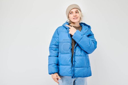 Téléchargez les photos : Jeune femme veste d'hiver bleue pointant du doigt, démontrant l'espace vide pour le produit ou le texte isolé sur fond de studio blanc - en image libre de droit