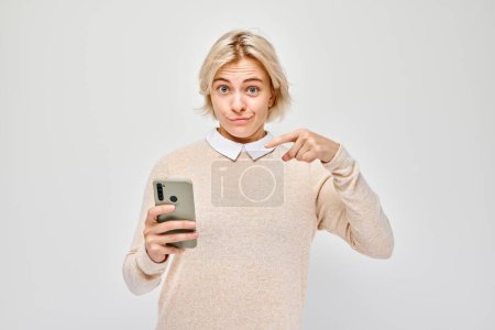 Foto de Retrato de una joven rubia mostrando la pantalla del teléfono móvil en blanco con la cara excitada. Persona con teléfono inteligente aislado en el fondo blanco - Imagen libre de derechos