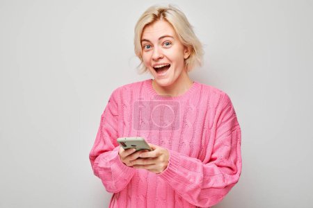 Foto de Retrato de una joven rubia en suéter rosa sosteniendo el teléfono móvil en la mano con la cara sonriente feliz. Persona con teléfono inteligente aislado en el fondo blanco - Imagen libre de derechos