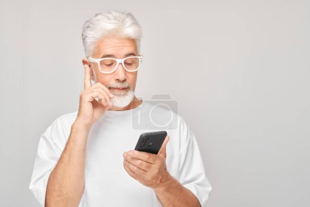 Téléchargez les photos : Portrait d'homme aux cheveux gris en T-shirt blanc regarde sur téléphone portable et pense. Personne avec smartphone isolé sur fond blanc - en image libre de droit