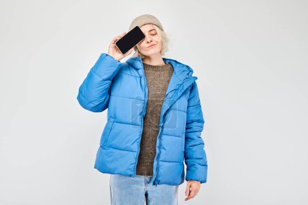 Foto de Retrato de una joven rubia con chaqueta azul que muestra la pantalla del teléfono móvil en blanco con la cara excitada. Persona con teléfono inteligente aislado en el fondo blanco - Imagen libre de derechos
