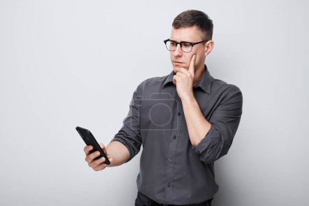 Foto de Retrato de hombre de negocios joven mira en el teléfono móvil y piensa. Persona con teléfono inteligente aislado en el fondo blanco - Imagen libre de derechos