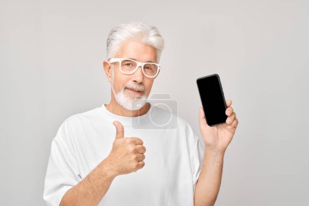 Foto de Retrato de hombre de pelo gris en camiseta blanca con teléfono móvil en la mano celebra la victoria, ganando dinero. Persona con teléfono inteligente aislado en el fondo blanco - Imagen libre de derechos