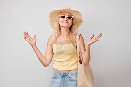 Foto de Retrato de mujer rubia madura con gafas de sol, sombrero, bolsa de compras celebrando vacaciones, saltando, bailando divirtiéndose aislado en fondo de estudio blanco - Imagen libre de derechos