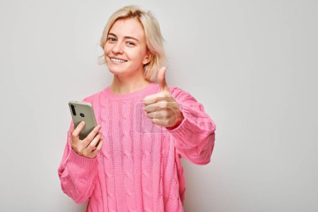 Photo for Portrait of young blond woman in pink sweater holding mobile phone in hand with happy smiling face. Person with smartphone isolated on white backgroun - Royalty Free Image