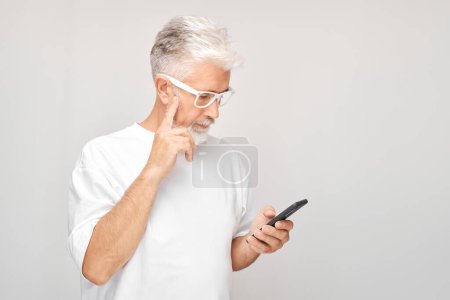 Foto de Retrato de hombre de pelo gris en camiseta blanca se ve en el teléfono móvil y piensa. Persona con teléfono inteligente aislado en el fondo blanco - Imagen libre de derechos