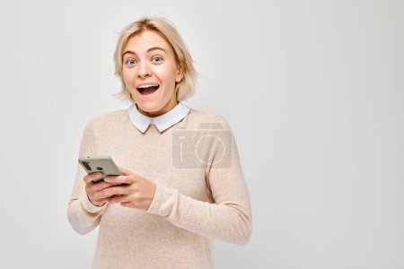 Téléchargez les photos : Portrait de jeune femme blonde tenant un téléphone portable à la main avec un visage souriant heureux. Personne avec smartphone isolé sur fond blanc - en image libre de droit