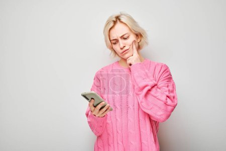 Photo for Portrait of young blond woman in pink sweater looks on mobile phone and thinks. Person with smartphone isolated on white background - Royalty Free Image