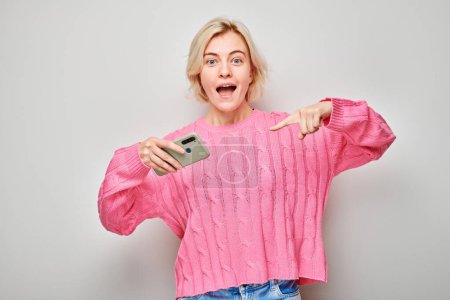 Foto de Retrato de una joven rubia en suéter rosa apunta con el dedo a la pantalla del teléfono móvil con la cara excitada. Persona con teléfono inteligente aislado en el fondo blanco - Imagen libre de derechos