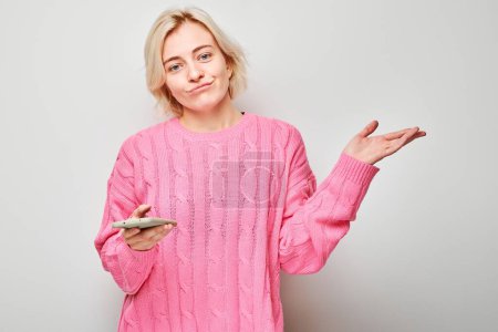 Téléchargez les photos : Portrait de jeune femme blonde en pull rose regarde sur téléphone portable et pense. Personne avec smartphone isolé sur fond blanc - en image libre de droit