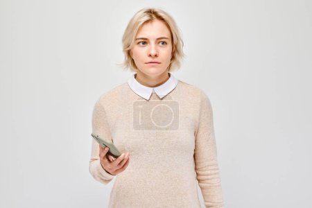 Foto de Retrato de mujer rubia joven mira en el teléfono móvil y piensa. Persona con teléfono inteligente aislado en el fondo blanco - Imagen libre de derechos