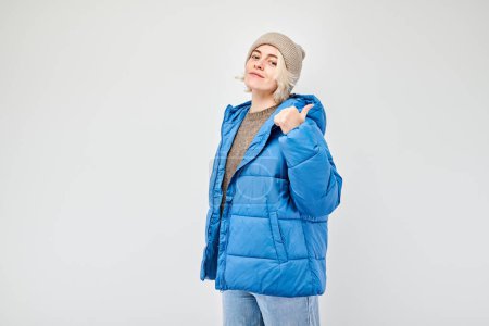 Foto de Mujer joven en chaqueta de invierno azul señalando a un lado el dedo, demostrando espacio vacío para el producto o texto aislado en fondo de estudio blanco - Imagen libre de derechos