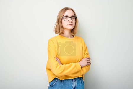 Foto de Mujer joven en suéter amarillo y gafas mirando hacia otro lado pensativamente sobre un fondo claro. - Imagen libre de derechos