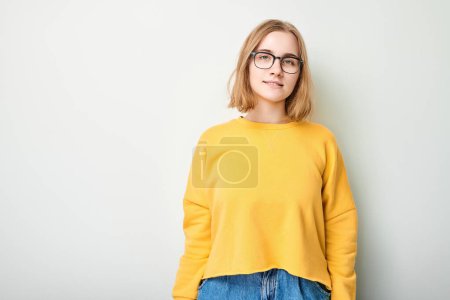 Téléchargez les photos : Jeune femme en pull jaune et lunettes debout sur fond clair avec un sourire confiant - en image libre de droit