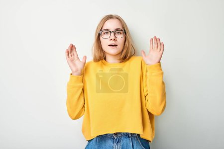 Young woman in yellow sweater throws up hands with shocked face, standing against white background