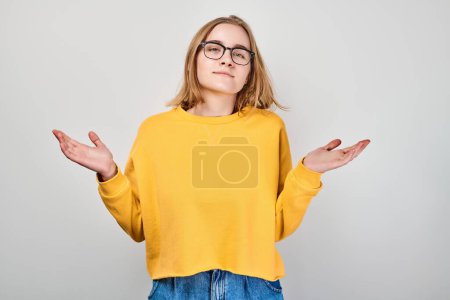 Foto de Mujer joven en suéter amarillo y gafas encogiéndose de hombros con expresión incierta sobre un fondo gris - Imagen libre de derechos