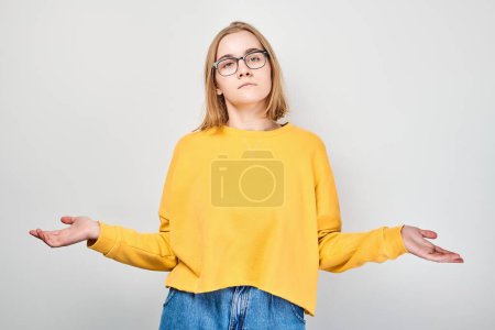 Téléchargez les photos : Jeune femme confus dans des lunettes et pull jaune haussant les épaules sur un fond gris. - en image libre de droit
