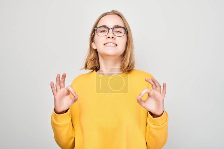 Photo for Smiling young woman in yellow sweater showing OK sign with both hands, isolated on grey background. - Royalty Free Image