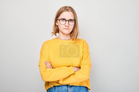 Téléchargez les photos : Jeune adulte confiant en pull jaune et lunettes debout avec les bras croisés sur un fond gris - en image libre de droit