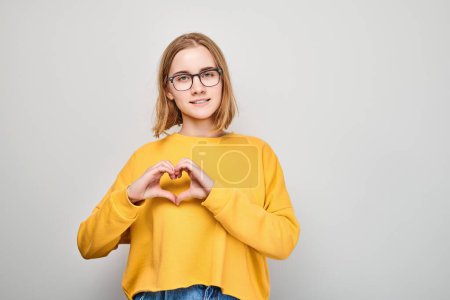 Foto de Mujer sonriente en suéter amarillo haciendo forma de corazón con las manos sobre fondo liso. - Imagen libre de derechos