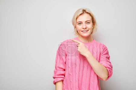 Téléchargez les photos : Jeune femme blonde en pull rose pointant du doigt, démontrant un espace vide pour le produit ou le texte isolé sur fond de studio blanc - en image libre de droit