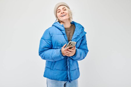 Téléchargez les photos : Portrait de jeune femme blonde vêtue d'une veste bleue tenant un téléphone portable à la main avec un visage souriant heureux. Personne avec smartphone isolé sur fond blanc - en image libre de droit