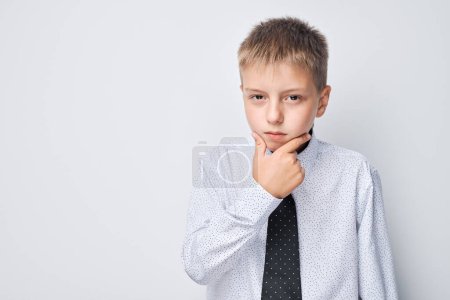 Foto de Niño pensativo en camisa y corbata con la mano en la barbilla sobre un fondo gris. - Imagen libre de derechos