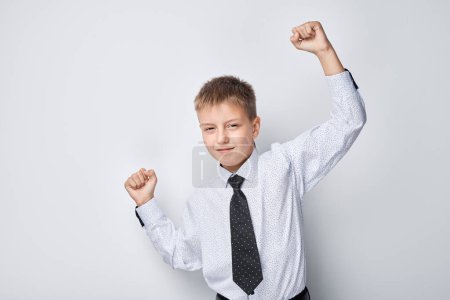 Téléchargez les photos : Joyeux jeune garçon en chemise et cravate célébrant le succès avec les poings levés sur un fond blanc - en image libre de droit