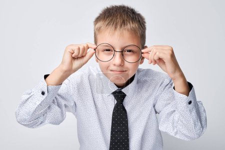 Téléchargez les photos : Jeune garçon en chemise et cravate faire un drôle de visage tout en ajustant les lunettes sur un fond clair. - en image libre de droit