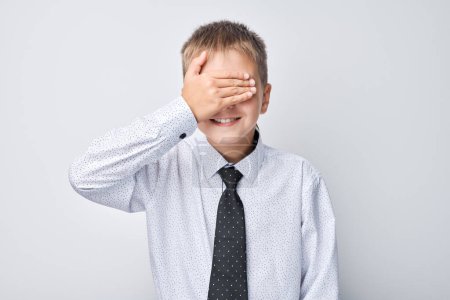 Photo for Smiling boy in shirt and tie covering his eyes with hand, playing hide and seek on plain background - Royalty Free Image