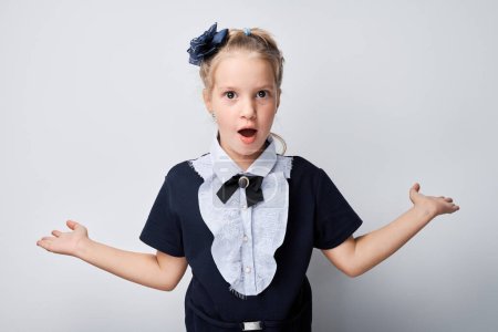 Photo for Surprised young girl with hands up, wearing bow and school uniform, isolated on light background. - Royalty Free Image
